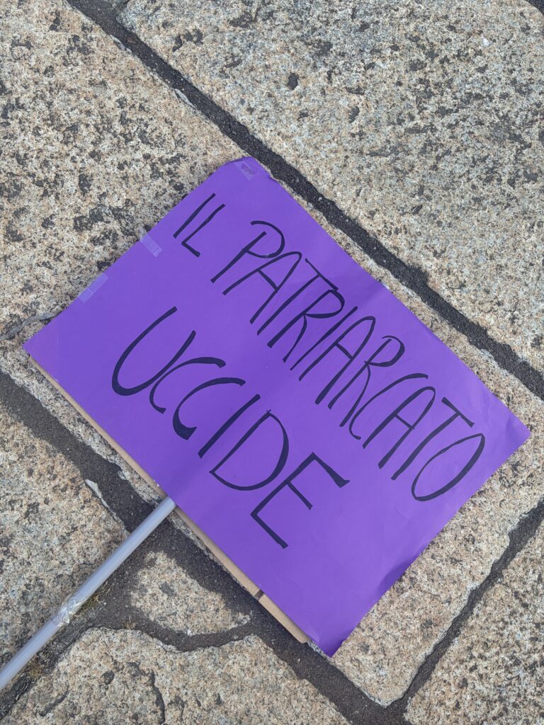 Cartello "Il patriarcato uccide": Manifestazione 8 marzo 2026, Casa delle donne Parma- Scritta viola su sfondo nero, su pavimento grigio, steso prima della partenza